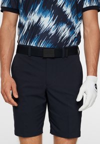 Helle marineblaue Golfsicht mit glatter Textur, mit einem schwarzen Gürtel und einem weißen Golfhandschuh an der rechten Hand. Darüber ein auffälliges Hemd mit blau-weißen Mustern.
