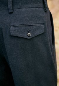 Dunkler, strukturierter Stoff der Hose, der eine Gesäßtasche mit einem Knopf zeigt, vor einem unscharfen neutralen Hintergrund.