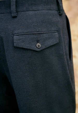 Mörk texturerad tyg av byxor, som visar en bakficka med en knapp, mot en suddig neutral bakgrund.