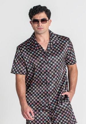 Mann trägt ein schwarzes kurzärmeliges Hemd mit Knöpfen und passende Shorts mit buntem "G"-Muster, Sonnenbrille, Hand in der Tasche, neutraler Hintergrund.