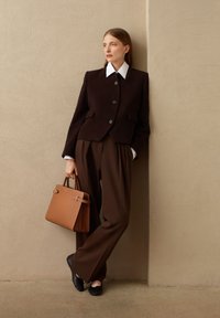 Blazer de lana marrón con botones y bolsillos negros, llevado sobre pantalones marrones de cintura alta. Bolso de mano de cuero marrón con detalles metálicos y zapatos planos.