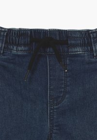 Pantalones de mezclilla azul oscuro con cintura elástica y cordón negro atado en un lazo en la parte frontal central.