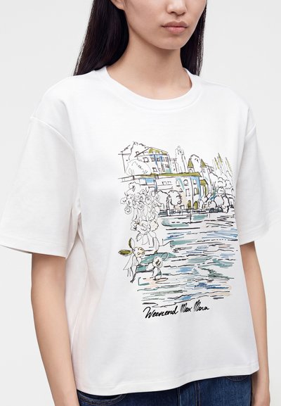 Γυναίκα που φοράει ένα λευκό T-shirt με ένα πολύχρωμο σκίτσο κτιρίων, δέντρων και νερού, υπογεγραμμένο "Weekend Max Mara" στο κάτω μέρος.