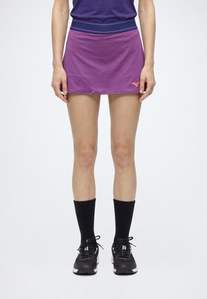 Un mannequin porte une jupe de sport violette avec une ceinture bleu marine, des chaussettes noires et des chaussures de course noires.