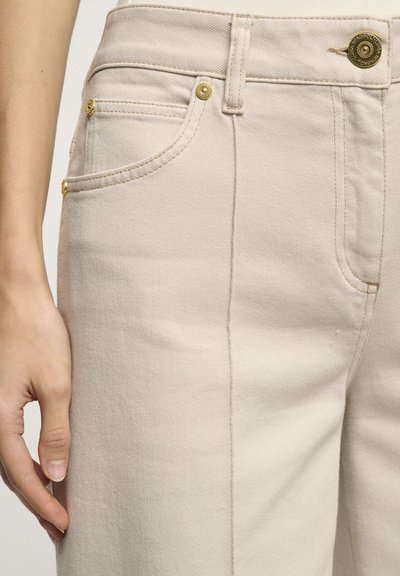 Beige denim hlače z gladko teksturo, opremljene z zlatimi gumbi, sprednjimi žepi in ravnim krojem.