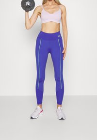 Žena v purpurových legínách a světle růžovém sportovním topu držící činku, stojící na jednoduché světle šedé podlaze a pozadí.