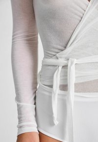 Biela dlhá rukávová blúzka s prekladaným dizajnom, vyrobená z priesvitnej látky a s viazaním v páse. Spodná časť je hladký biely kúsok, zdôrazňujúci kontrast textúr.