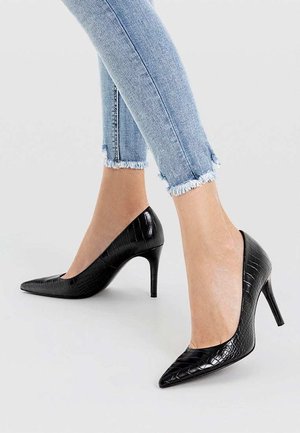 Pieds portant des escarpins noirs à bout pointu avec un motif crocodile, associés à un jean skinny bleu clair à ourlet effiloché sur fond blanc.