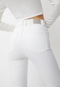 Žena v bílých džínách s vysokým pasem a bílém dlouhém cropped topu, ukazující zadní kapsy a štítek na pase.