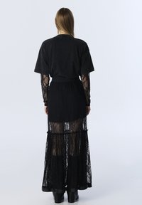 Zwarte jurk met een doorschijnende kanten overlay, lange mouwen, gelaagd ontwerp en een aansluitende tailleband. Zwarte enkellaarsjes maken de look compleet.