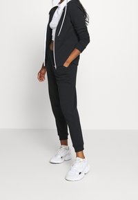 Felpa nera con zip e cordini bianchi, abbinata a pantaloni jogger coordinati e sneakers bianche. Vestibilità casual con tasche sulla felpa e sui pantaloni.
