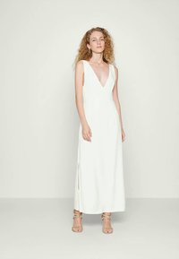 Vestido maxi branco sem mangas com um decote em V profundo, fendas laterais e textura suave. A modelo também usa sandálias com tiras e contas.
