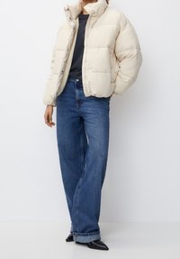Giacca a vento leggera beige, con colletto alto e bottoni a pressione. Abbinata a pantaloni in denim blu larghi e scarpe nere appuntite.