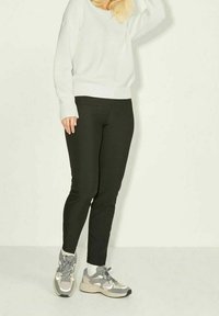 Personne portant un pull blanc, un pantalon slim noir, des chaussettes blanches et des baskets Nike grises, debout devant un fond clair uni.