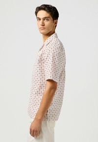 Camicia a maniche corte di un rosa chiaro con un motivo floreale ripetuto in bordeaux. Design con colletto e patta con bottoni, realizzata in tessuto leggero.