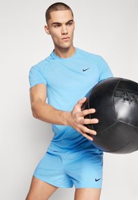Lys blå atletisk skjorte med en svart Nike-logo, kombinert med matchende shorts. Modellen holder en svart medisinball, og viser frem et tett design.