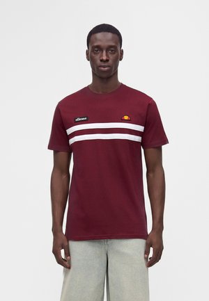 Uomo che indossa una maglietta color bordeaux con due strisce orizzontali bianche e loghi sul petto, abbinata a pantaloni grigio chiaro.