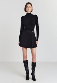 Maglione a collo alto nero abbinato a una mini gonna plissettata nera con dettagli in metallo. Modella con stivali neri con tacco fino al ginocchio.