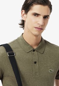Polo de color verde oliva con textura, con cuello, dos botones negros y un pequeño logo bordado. Tirante negro en el hombro.
