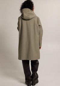 Pitkä beige sadetakki huppuineen, valmistettu sileästä, vedenpitävästä materiaalista. Yhdistetty löysiin tummanharmaisiin housuihin ja mustiin saappaisiin.