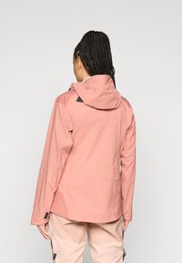 Casaco impermeável rosa suave com capuz, apresentando detalhes pretos nos ombros e tecido texturizado. Costuras visíveis e corte descontraído.