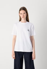 White cotton t-shirt with short sleeves, round neck, and a small black "CK" logo on the left chest. Paired with dark denim pants.