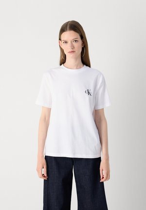 White cotton t-shirt with short sleeves, round neck, and a small black "CK" logo on the left chest. Paired with dark denim pants.