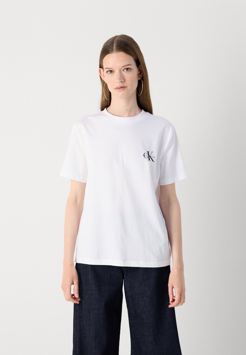 White cotton t-shirt with short sleeves, round neck, and a small black "CK" logo on the left chest. Paired with dark denim pants.
