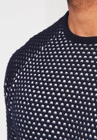 Pull en tricot bleu présentant un motif géométrique texturé, col rond et poignets côtelés. Le matériau semble léger et respirant.