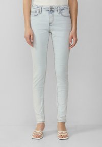 Jean skinny bleu clair avec une texture denim lisse, design à cinq poches et un subtil détail de couture le long des coutures. Associé à des sandales blanches à lanières.