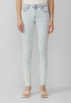 Helles blaues Skinny-Jeans mit glatter Denim-Oberfläche, Fünf-Taschen-Design und dezentem Steppdetail entlang der Nähte. Kombiniert mit weißen Riemchensandalen.