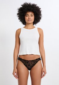 Top corto in cotone bianco con spalline larghe, abbinato a un perizoma in pizzo nero con motivi floreali e un accento rosa al centro.