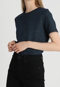 Mørkeblå kortærmet t-shirt med en diskret glans, med rund halsudskæring og en tætsiddende pasform, kombineret med sorte jeans.