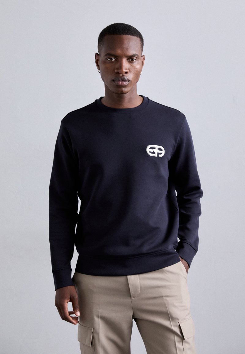 Tengerészkék kapucnis pulóver kerek nyakkal és hosszú ujjakkal. Elölnézetén fehér logó található, bordázott mandzsettákkal és aljjal. Sima tapintású.