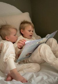 Kaksi lasta vaaleanbeigeissä kukkakuosisissa pyjamissa istuu sängyssä, luettuna intensiivisesti värikästä satukirjaa yhdessä.
