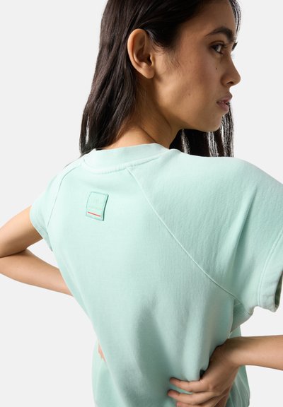 Junge Frau mit langem dunklem Haar trägt ein helltürkisfarbenes Kurzarmshirt, Hände in die Hüften gestützt, seitlich stehend vor einfachem weißem Hintergrund.