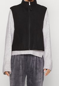 Svart ärmlös väst med hög krage och framdragkedja, bärs över en grå långärmad topp. Har en kortad design och mjuk tygtextur.