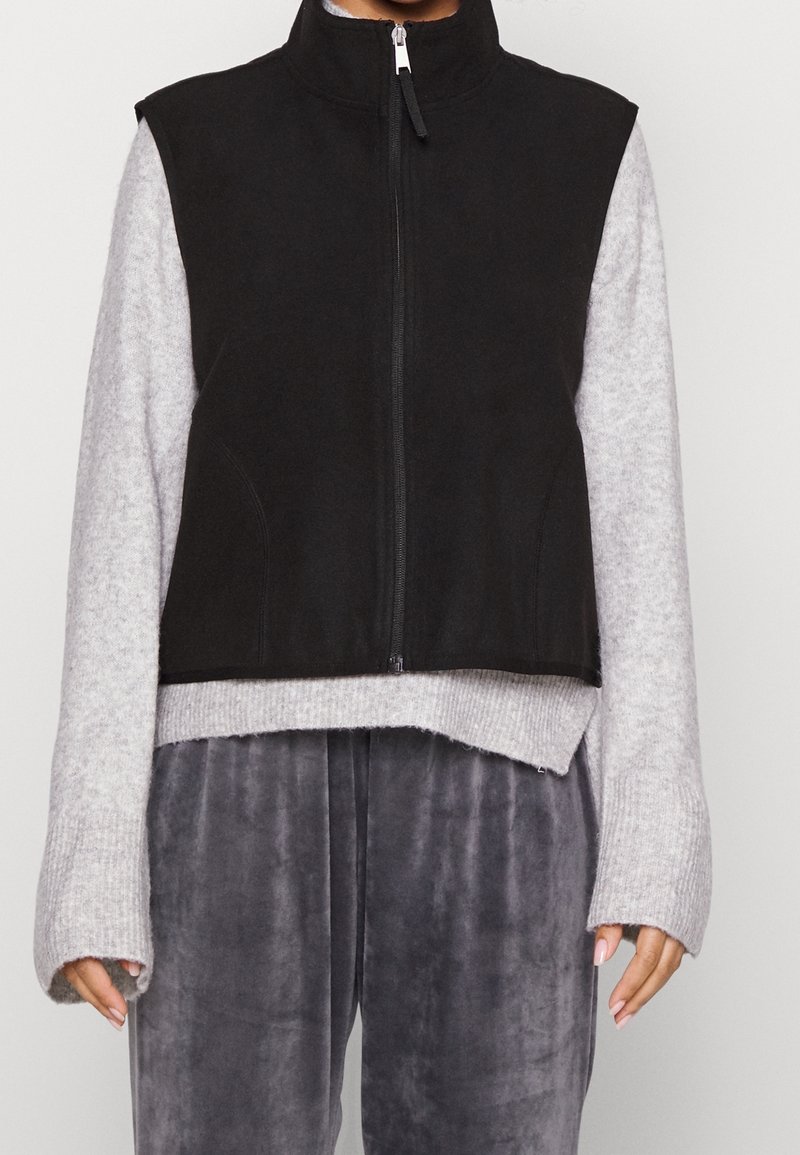 Svart ärmlös väst med hög krage och framdragkedja, bärs över en grå långärmad topp. Har en kortad design och mjuk tygtextur.