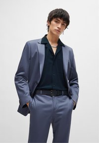 Un costume slim-fit bleu clair avec des revers aigus. Porté sur une chemise sombre et texturée. Présente une ceinture noire et des détails en métal argenté.