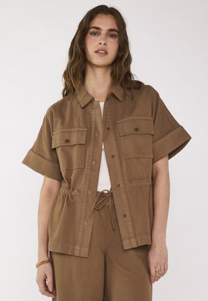 Vrouw draagt een losse bruine korte mouwen jas met borstzakken over een witte top en bijpassende bruine broek met een trekkoord in de taille.