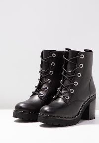Botas de cuero negras hasta el tobillo con tacones gruesos, parte delantera con cordones y ojales de metal, y detalles de tachuelas a lo largo de los bordes de la suela sobre una superficie blanca.