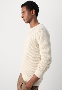 Pull en maille beige clair avec une texture en tricot ouvert, col rond et poignets côtelés, associé à un pantalon beige.