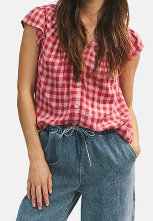 Frau trägt eine rot-weiß karierte Bluse mit Knöpfen und Rüschensäumen sowie blaue Jeans mit Tunnelzug, eine Hand in der Tasche.