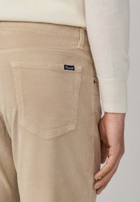 Beige Corduroyhosen mit Gesäßtasche und kleinem schwarzen Stoffetikett, getragen mit einem cremefarbigen Langarmpullover, Hand entspannt an der Seite.