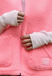 Roza flis telovnik z teksturirano površino, z zadrgo spredaj in dvema žepoma spredaj. Bela dolga majica z luknjicami za palce.