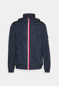 Giacca leggera navy con colletto alto, zip rossa e bianca, polsini elasticizzati e una leggera lucentezza; include una tasca laterale con pattina.
