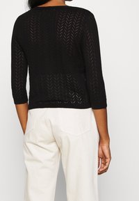 Suéter negro de punto con un patrón texturizado en zigzag, mangas de tres cuartos y un ajuste relajado, combinado con pantalones de color claro.