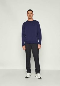 Μπλούζα σε ναυτικό μπλε χρώμα με μακρυά μανίκια, με ραμμένα μανίκια και μέση, σε συνδυασμό με σκούρες παντελόνες και λευκά αθλητικά παπούτσια. Ορατή διακριτική λεπτομέρεια λογότυπου.