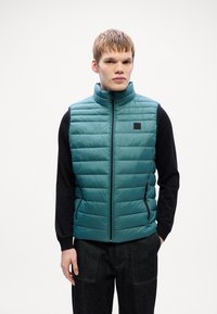 Gilet matelassé en teal avec col haut, fermeture éclair et poches latérales. Surface texturée avec un patch de marque noir sur la poitrine.