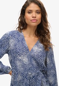 Vestido envolvente azul con patrón floral blanco, mangas largas, escote en forma de v y detalle de cintura con lazo, fabricado en una tela ligera con una textura suave.